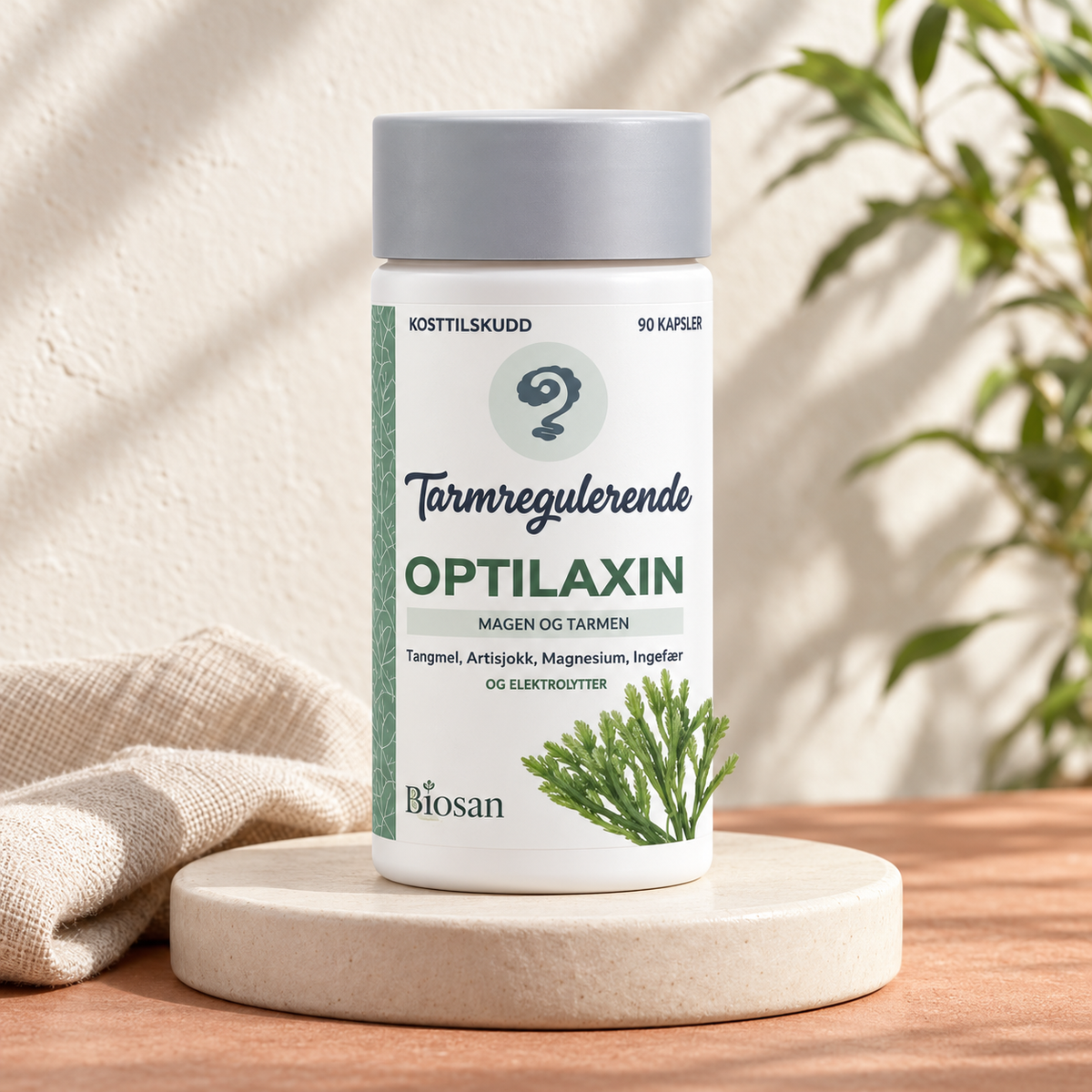Biosan Optilaxin 90 kapsler på en lys keramisk sokkel med lintekstil og grønne planter i bakgrunnen.